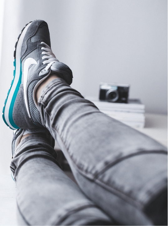 A person wearing jeans and athletic shoes. Their legs are crossed on a desk